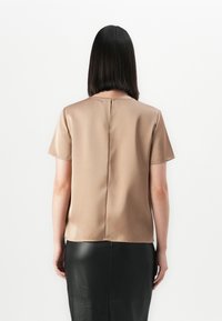 Vrouw met steil, donker haar, gekleed in een beige blouse met korte mouwen en een zwarte leren rok, staand met haar gezicht naar een witte achtergrond.