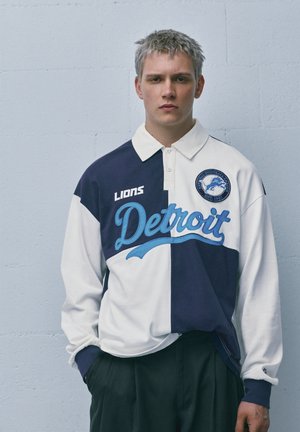 Mladý muž s krátkymi blond vlasmi v tmavomodro-bielom rugby tričku tímu Detroit Lions, stojaci pred jednoduchou svetlomodrou stenou.