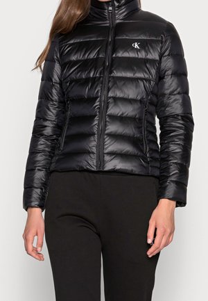 Vrouw draagt een aangesloten zwarte pufferjas met rits aan de voorkant en het Calvin Klein-logo op de borst, gecombineerd met een zwarte broek.