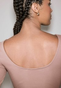 Long-sleeve top in light pink ribbed fabric, featuring a wide scoop back neckline and smooth texture, with hair styled in braids.