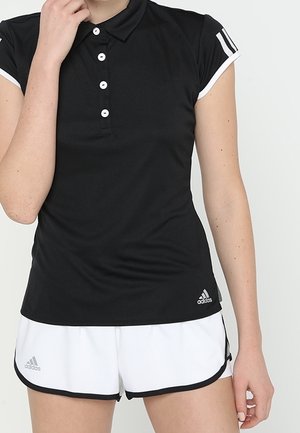 Femme portant une chemise Adidas noire à col avec des boutons blancs et un short de sport Adidas blanc avec une bordure noire.