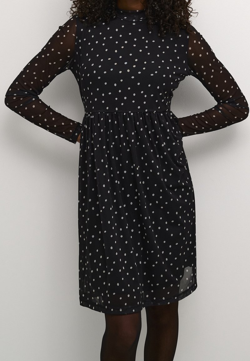 Femme portant une robe noire à manches longues avec des pois blancs et des manches transparentes, debout les mains sur les hanches devant un fond uni.