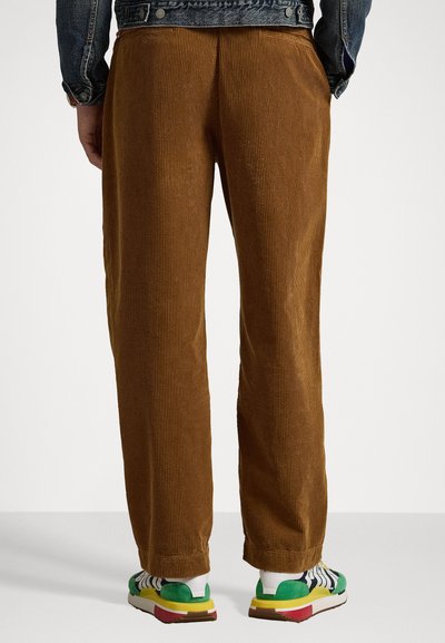 Pantaloni din catifea maro cu croială dreaptă, prezentând ribbing vertical și un croi confortabil, asortați cu adidași multicolori.