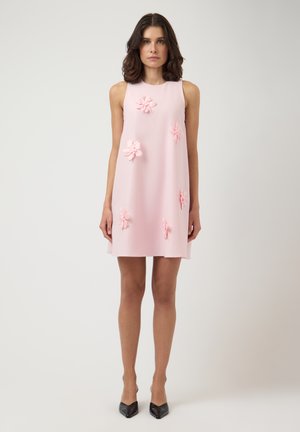 Vrouw draagt een mouwloze lichtroze A-lijn jurk met 3D bloemdetails, gecombineerd met zwarte pumps met spitse neus, staand tegen een effen achtergrond.