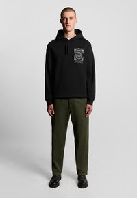 Felpa nera con grafica frontale, tasca a marsupio, cappuccio con cordino; abbinata a pantaloni verde oliva e scarpe nere con suola spessa.