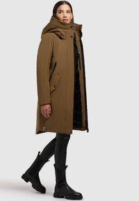 Parka marron avec une capuche, fermeture éclair à l'avant et fermeture à pression, deux poches latérales, doublure noire visible, porté sur un pantalon noir ajusté et des bottes.