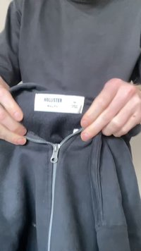 Osoba trzyma ciemnoszarą bluzę rozpinaną z kapturem, pokazując metkę marki "Hollister" wewnątrz kołnierza z rozmiarem medium oraz szczegółami dotyczącymi dopasowania.