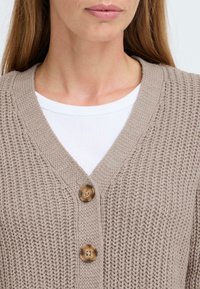 Beige knitted cardigan with a V-neck, featuring two large, round buttons. A white ribbed shirt is visible beneath the cardigan.