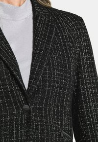 Schwarzer und weißer strukturierter Blazer mit Reverskragen, einreihiger schwarzer Knopfverschluss und seitlicher Reißverschlusstaschen. Getragen über einem hellgrauen Rollkragenpullover.