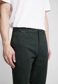 Tmavě zelené kalhoty z bavlny, s plochým předním dílem, poutky na opasek a zapínáním na jeden knoflík. Nenápadné značení viditelné v pase.