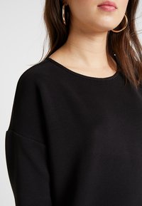 Musta ribatusta neuleesta, jossa on leveä, pyöreä kaula-aukko ja alas lasketut olkapäät. Silkkinen pinta, jossa on hienovaraisia vaakasuoria viivoja. Suuret renkaat korvakorut näkyvissä.