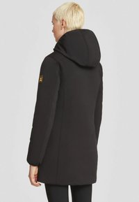 Cappotto imbottito nero con cappuccio, texture liscia, vestibilità slim. Nota patch logo giallo sulla manica, cuciture piatte e design minimalista.