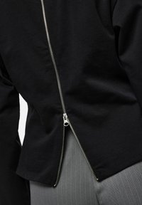 Haut noir à manches longues avec un détail de fermeture éclair argentée le long de la couture arrière, fabriqué en coton, offrant une coupe décontractée et une texture lisse.