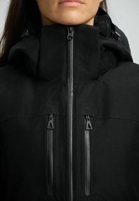 Schwarze isolierte Jacke mit hohem Kragen, ausgestattet mit sichtbaren Reißverschlüssen und Kordeln, aus einem weichen, strukturierten Stoff gefertigt.