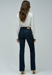 Jeans de mezclilla de un azul oscuro con una cintura alta, diseño acampanado y bolsillos traseros. Combinados con una blusa blanca abotonada y tacones nude.