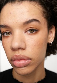 Gros plan d'un visage avec une peau brun moyen, des cheveux courts et ondulés, des yeux marron et une couleur de lèvres naturelle. Les traits comprennent la texture de la peau et de légères taches de rousseur.