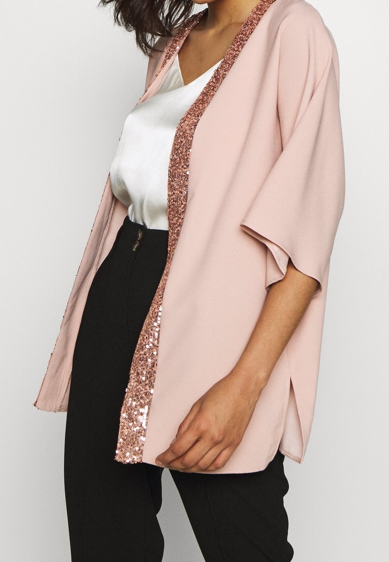 Vaaleanpunainen takki, joka on valmistettu pehmeästä kankaasta ja jossa on löysä istuvuus. Edessä ja hihoissa on kimalteleva ruusunkultainen paljettireuna.