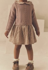 Pull en tricot rose avec un col et un motif texturé, associé à une jupe crème à motifs floraux et des chaussures marron avec des chaussettes volantes.