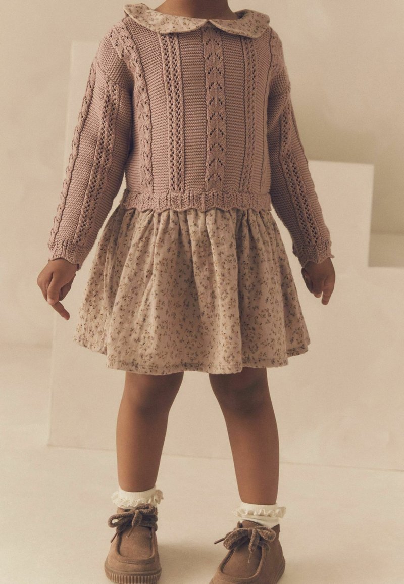 Pull en tricot rose avec un col et un motif texturé, associé à une jupe crème à motifs floraux et des chaussures marron avec des chaussettes volantes.