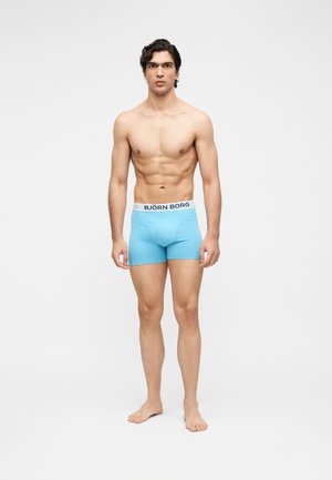 Muskløs mann står barfot og har på seg lyseblå Björn Borg boxershorts foran en ensfarget hvit bakgrunn.