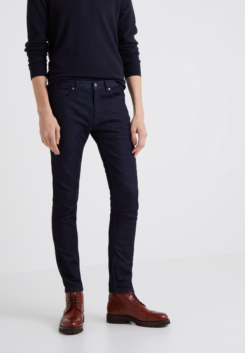 Mörkblå slim-fit jeans i denim, med en klassisk femficksdesign och metallhardware. Kombinerade med bruna snörstövlar.