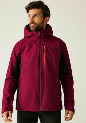 Mann trägt eine bordeauxrote und schwarze Outdoor-Jacke mit Kapuze und Reißverschlusstasche auf der Brust, schaut leicht nach links.