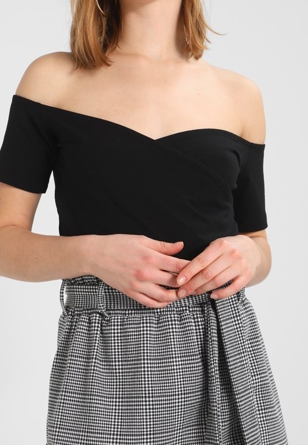 Femme portant un haut noir épaules dénudées et une jupe à carreaux noire et blanche taille haute, debout les mains doucement jointes.