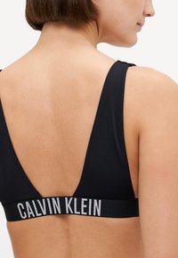 Fekete sportmelltartó mély V-hátrésszel, széles pántokkal és puha elasztikus pánttal, amelyen fehér "CALVIN KLEIN" felirat található. Simább anyagú textúra.