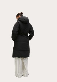 Abrigo largo acolchado negro con capucha, que presenta una cintura ajustada y una textura suave, acompañado de pantalones de pierna ancha a rayas blancas.