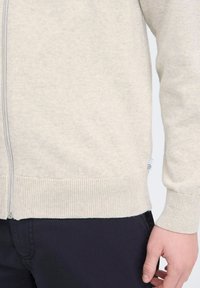Sudadera cremoso de cierre con cremallera, hecha de un tejido suave. Presenta puños y dobladillo acanalados, con una pequeña etiqueta de logo en el lado.