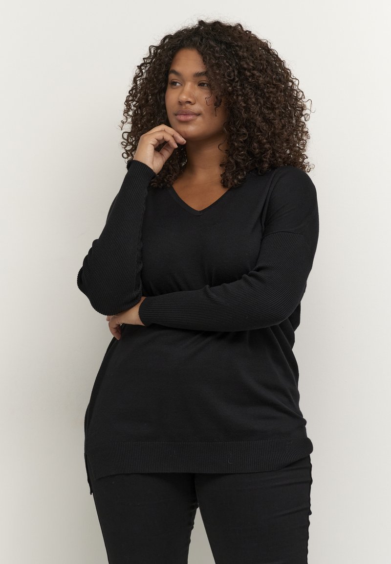 Schwarzer Strickpullover mit V-Ausschnitt, gerippten Ärmeln und seitlichen Schlitzen. Tragen mit schwarzen Hosen, die einen lässigen Schnitt und eine glatte Textur aufweisen.
