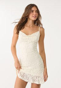 White lace mini dress with a fitted design, spaghetti straps, and a ruffled neckline detail. Textured lace overlay adds dimension.