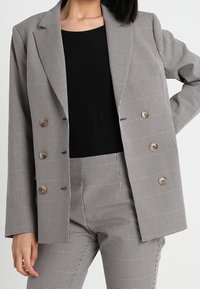 Šedý kostkovaný blazer se strukturovaným designem, dvouřadým zapínáním a hnědými knoflíky, vrstvený přes černý top a odpovídající kalhoty.