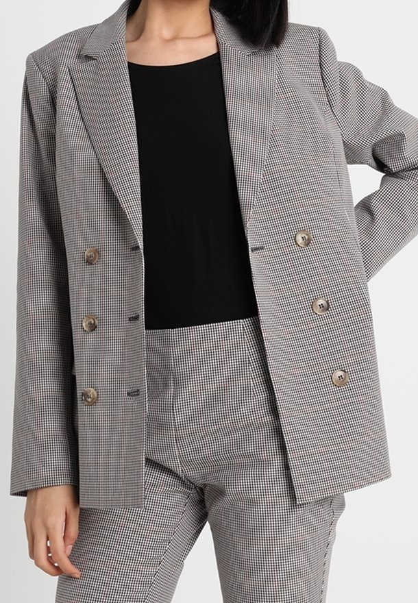 Šedý kostkovaný blazer se strukturovaným designem, dvouřadým zapínáním a hnědými knoflíky, vrstvený přes černý top a odpovídající kalhoty.