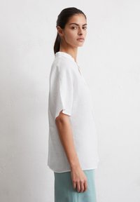 Weißes Leinenhemd mit kurzen Ärmeln und Stehkragen, entspannter Schnitt und subtiler Struktur. Getragen mit einem hellblauen Rock.