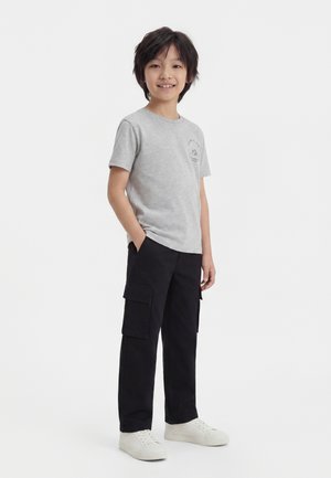 Dreng med sort hår iført en lysegrå kortærmet t-shirt, sorte cargobukser og hvide sneakers, der står med hænderne i lommerne.