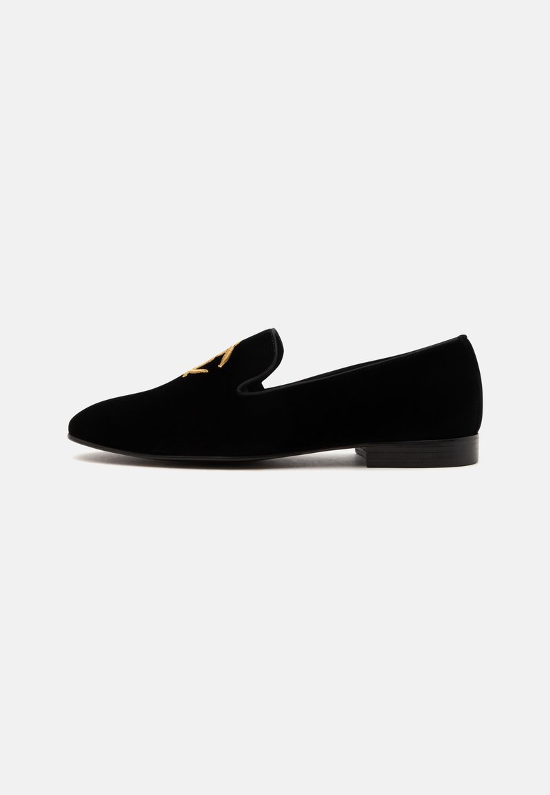 Μαύρο βελούδινο παπούτσι slip-on με χαμηλό τακούνι και χρυσή κεντημένη λεπτομέρεια στο επάνω μέρος, απεικονίζεται σε προφίλ πλαγίως σε λευκό φόντο.