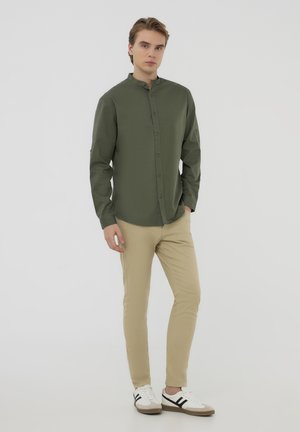 Camicia verde a maniche lunghe con colletto a banda, chiusura a bottoni e texture liscia, abbinata a pantaloni slim-fit beige e scarpe bianche casual.