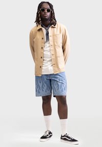 Chaqueta caqui con botones, camiseta blanca, pantalones cortos de mezclilla azules con un patrón de llamas, zapatillas negras con acentos blancos y gafas de sol.