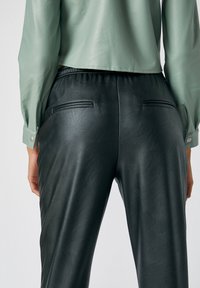 Des pantalons en simili cuir noir présentent une texture lisse, une coupe ajustée et des poches arrière. Une veste en cuir vert clair complète l'ensemble.