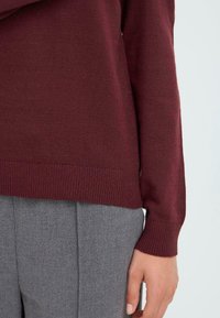 Burgundowa dzianinowa sweter z prążkowanymi mankietami i dołem, zestawiony z szarymi teksturowanymi spodniami, podkreślający detale materiału i kontrast kolorów.