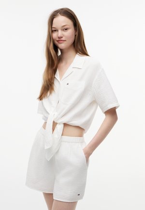 Ung kvinde med langt hår iført en hvid kortærmet skjorte bundet i taljen og matchende hvide strukturerede shorts med lommer.