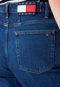 Mørkeblå denimjeans har en rektangulær lapp med "TOMMY JEANS" og en rød, hvit og blå logo over baklommen.
