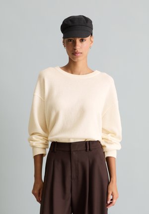 Femme portant un pull crème à manches longues, un pantalon taille haute marron foncé et une casquette plate noire, debout devant un fond gris uni.