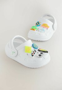 Sabots blancs ornés de charms et d'autocollants colorés et ludiques, présentant divers motifs et motifs vibrants pour un look amusant.