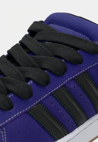 Μωβ σουέτ αθλητικό παπούτσι με μαύρα κορδόνια και τρεις μαύρες ρίγες. Διαθέτει λευκή ραφή και λαστιχένια σόλα, με υφή επιφάνειας.