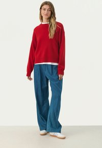 Red oversized sweater with a crew neck, layered over a white shirt, paired with loose blue denim pants and white shoes.