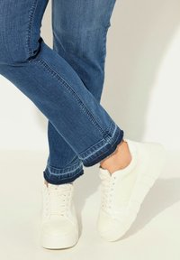 Bílé šněrovací tenisky s texturovaným povrchem, spárované s modrými džíny s třepenými okraji a úzkým střihem.