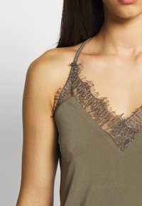 Camiseta sin mangas de color verde olivo con un borde de encaje a lo largo del escote y los hombros, con un delicado detalle de flecos y tirantes finos.