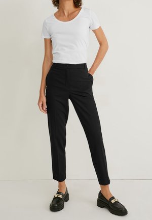 Femme portant un t-shirt blanc à manches courtes, un pantalon noir ajusté aux chevilles et des mocassins noirs avec une boucle dorée, se tenant devant un mur uni.